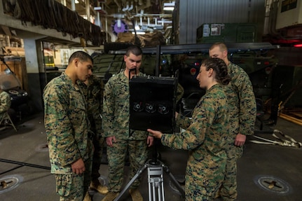 Three men and a woman wearing camouflage military uniforms examine a piece of military equipment while aboard a ship. There is additional military equipment in the background.