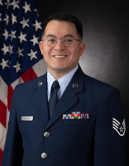 Official Photo of SSgt Christopher Arellano, Guitarist, United States Air Force Bands. SSgt Arellano is wearing navy blue service dress in front of an American flag.