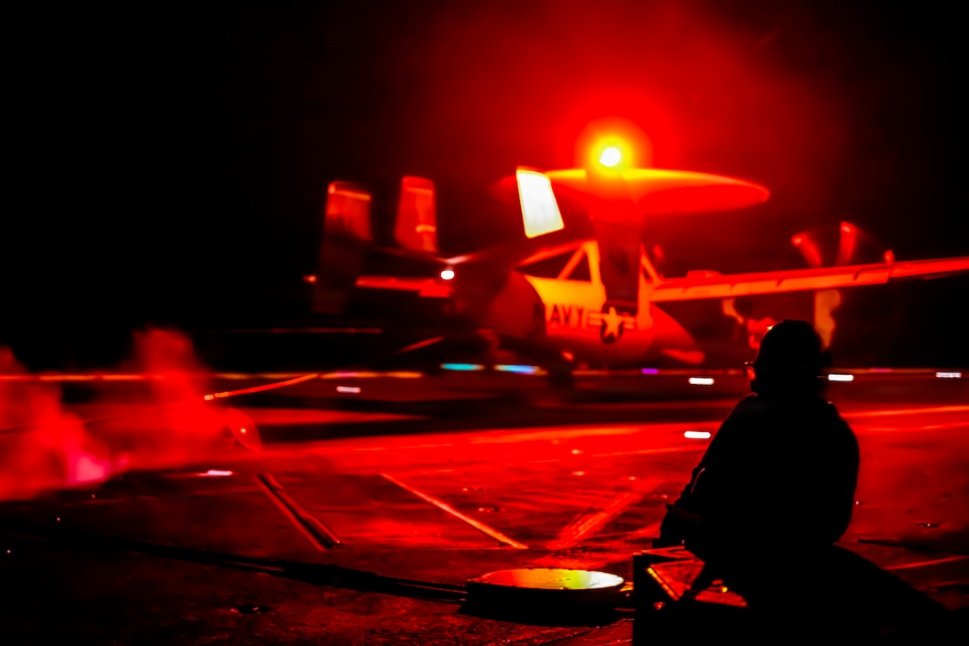 A kneeling sailor, shown from behind in silhouette, faces an aircraft on a ship's deck against a dark sky illuminated by red light.