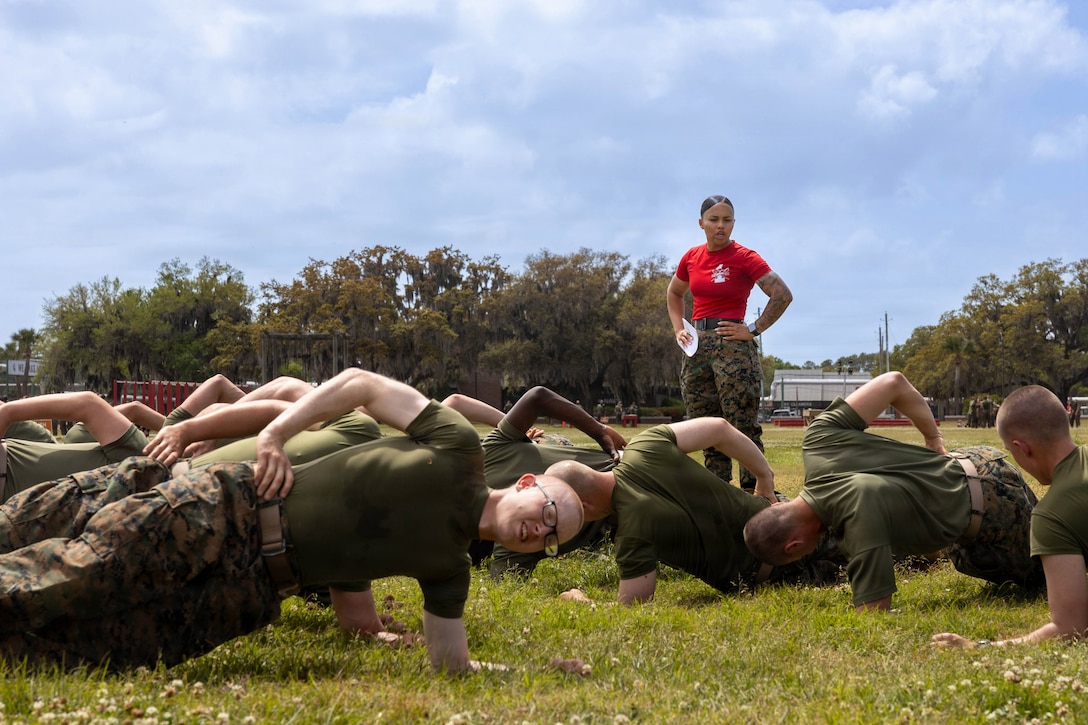 A Marine in a red T-shirt stands and watches fellow Marines holding themselves in plank positions in a grassy field.