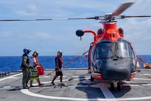 A man and a woman in casual attire and life jackets are escorted by a man in a military flight suit and aviation headgear onto a military helicopter on a large ship.