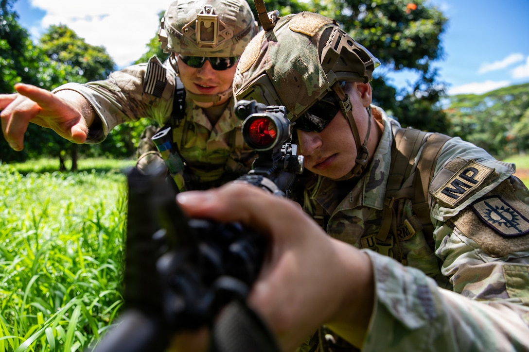 A soldier aims a weapon as another points in the same direction in a grassy area.