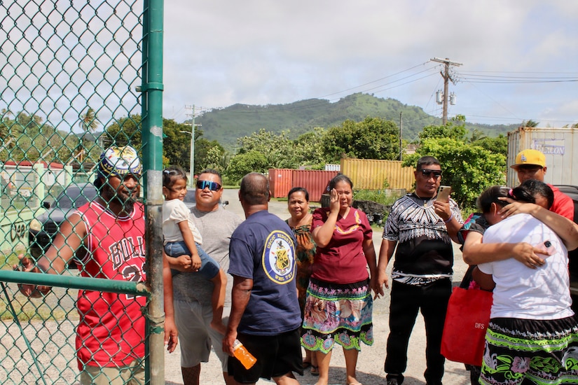 Two women in casual attire embrace outside a gate as eight other people in similar attire stand beside them.