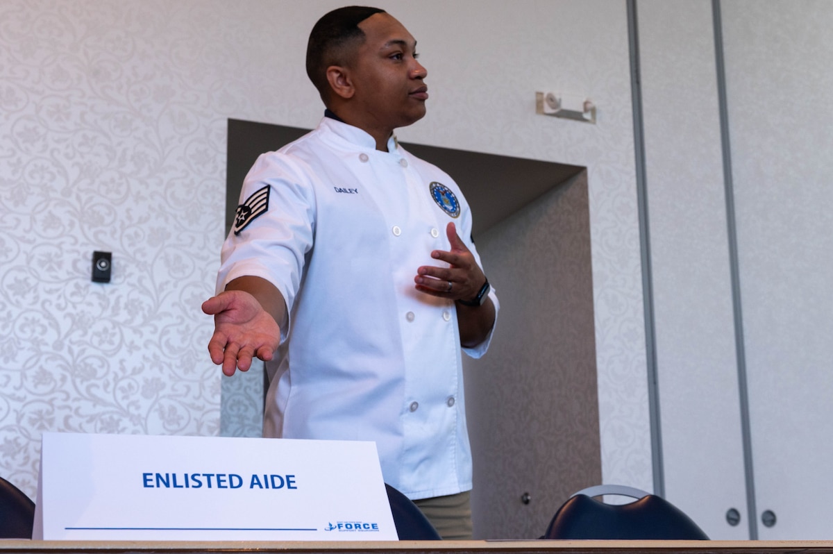 A gentleman in a white coat stands with his arm towards a card. The card reads "Enlisted Aide."