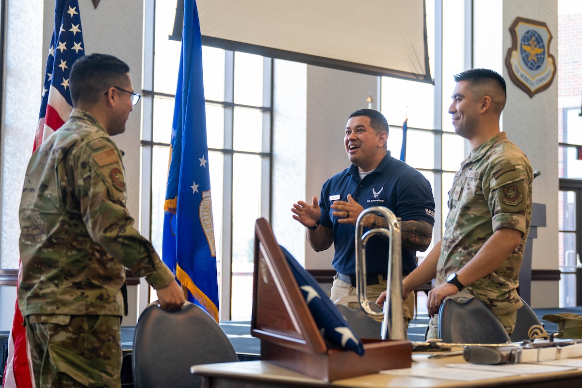 3 gentlemen stand behind a table that has a US flag and a bugle. Man to the left looks towards 2 men on the right. The men in the center has his hands up, with a smile, and the man on the right is smiling with his hands in front of himself.
