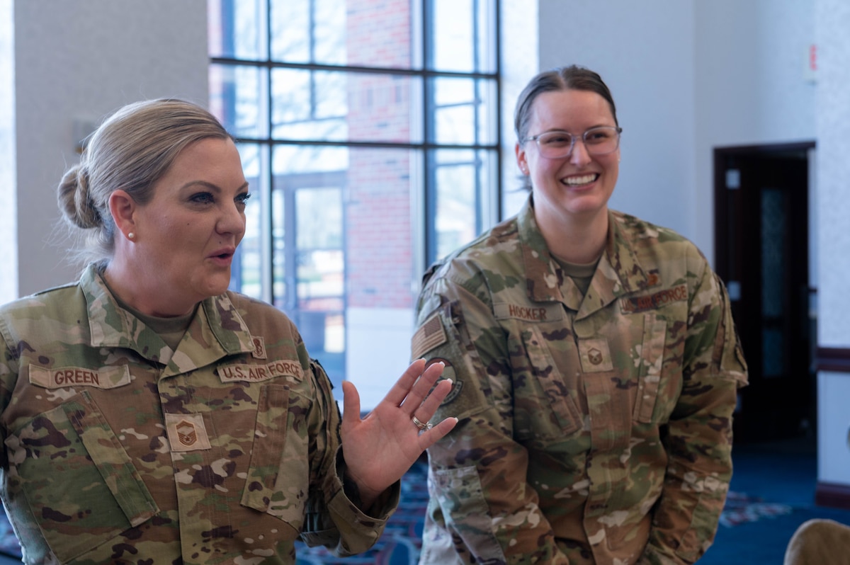 2 ladies stand, talking to someone off the photo. The lady on the left has her hand up, as if she is directing someone in a joking manner, and the lady to the right has a smile, laughing along with the conversation.