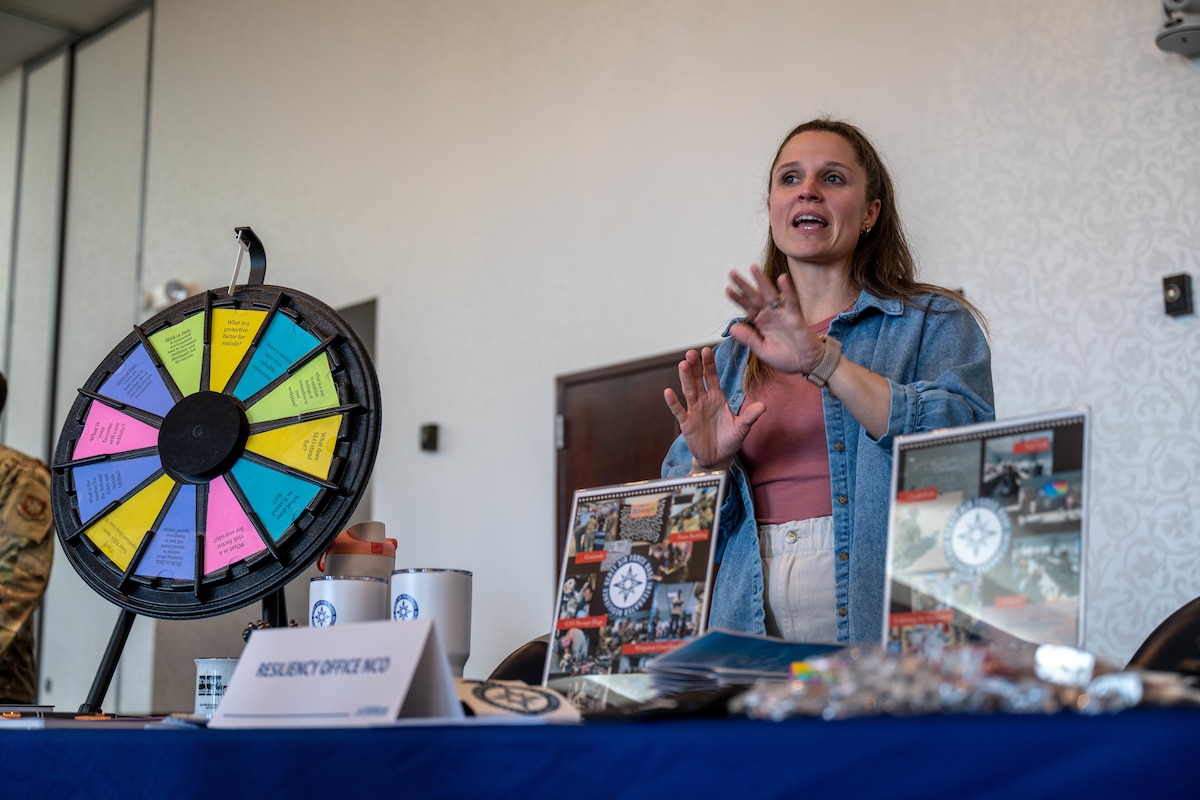 A woman stands behind a table with items laid out, displaying her career field. She is speaking to someone who is out of the photo.
