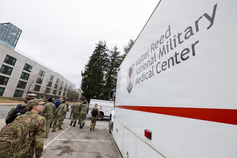A group of people dressed in camouflage military uniforms walk in line next to an emergency vehicle that has the words "Walter Reed National Military Medical Center" on it.
