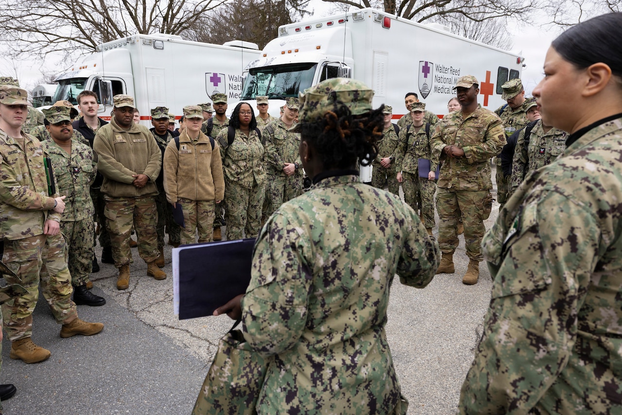 People in camouflage military uniforms look at a woman in similar attire at the center of the crowd while she holds up a notebook. Emergency vehicles are parked in the background.