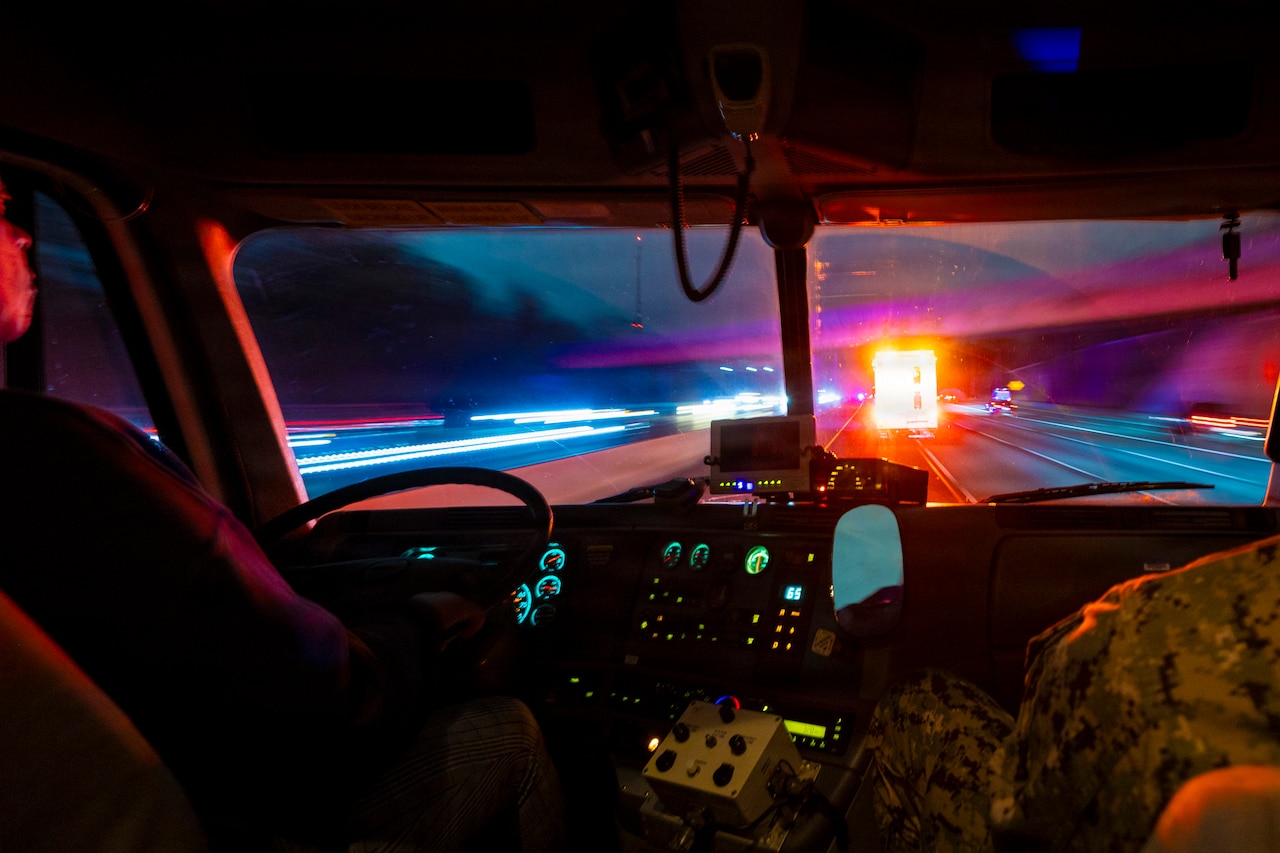 A person in a camouflage military uniform drives an emergency vehicle at night while another, partially visible person in similar attire sits in the passenger seat.