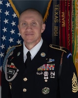 Army Soldier in dress uniform with yellow command sergeant major stripes on his sleave and various ribbons and badges on both sides of his jacket. In the background is the U.S. Flag on the left and the Army flag with streamers visible on the right.