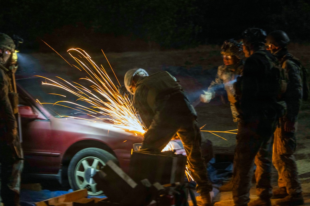 Sparks fly against a dark sky as a Marine uses a tool to cut into a car's hood while fellow Marines watch.