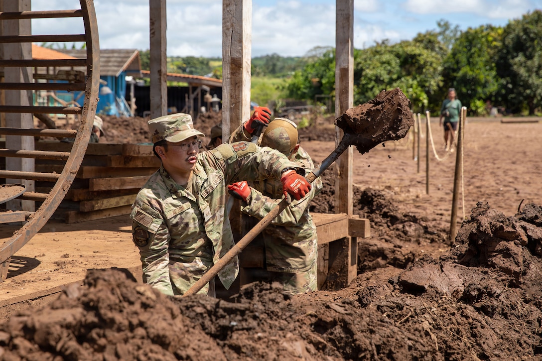 Two airmen use shovels while standing knee-deep in dirt by a wooden structure.