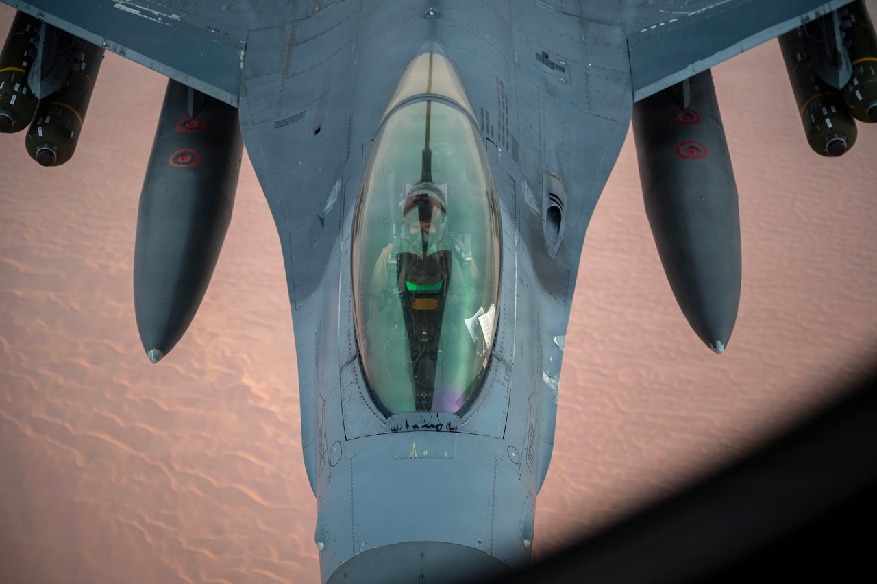 A pilot is visible inside the cockpit of a fighter aircraft seen from above as it flies over desert-type terrain.