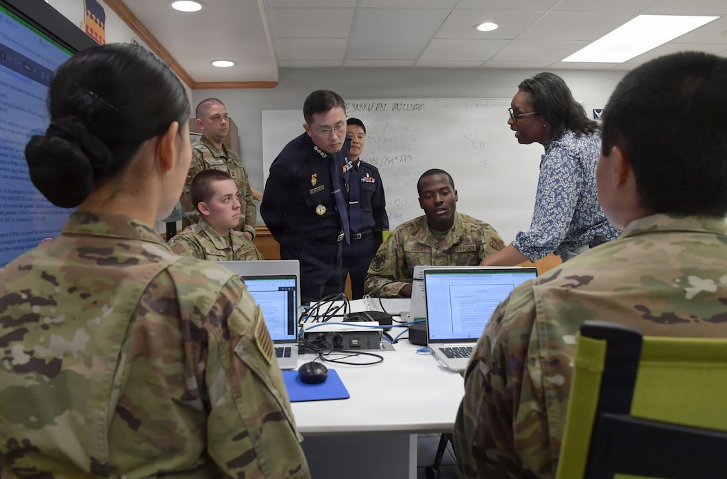 A group of people in military uniforms stand and sit while staring at computer screens.