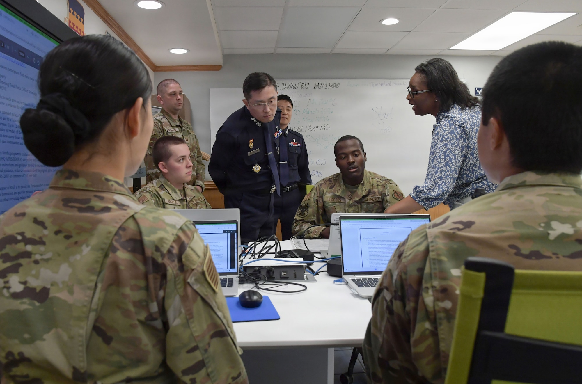 A group of people in military uniforms stand and sit while staring at computer screens.