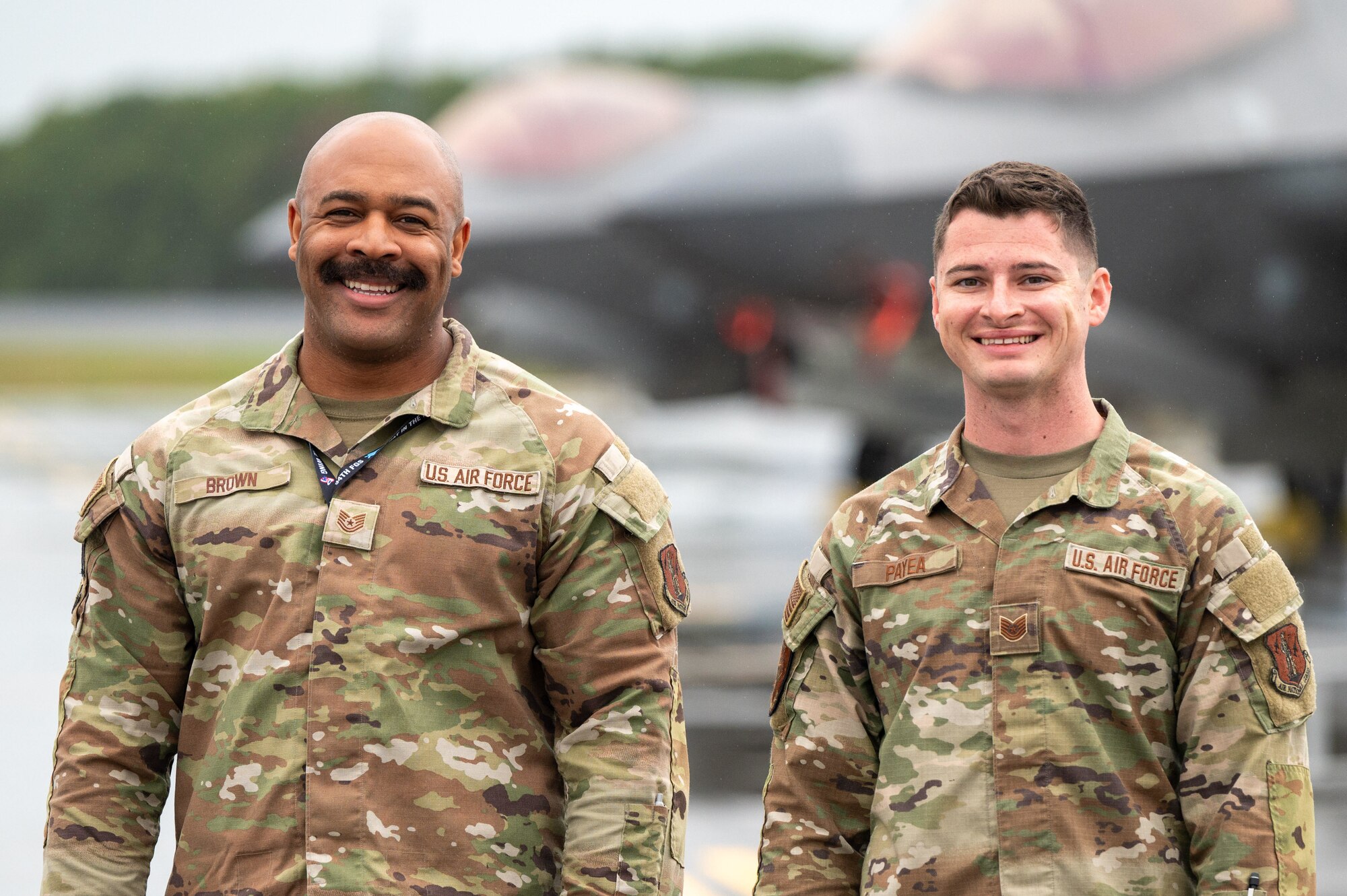 Two U.S. Air Force service members in OCP uniforms smile for a photo on an airfield, with the blurred grey shape of an F-35 Lightning II stealth fighter jet behind them.