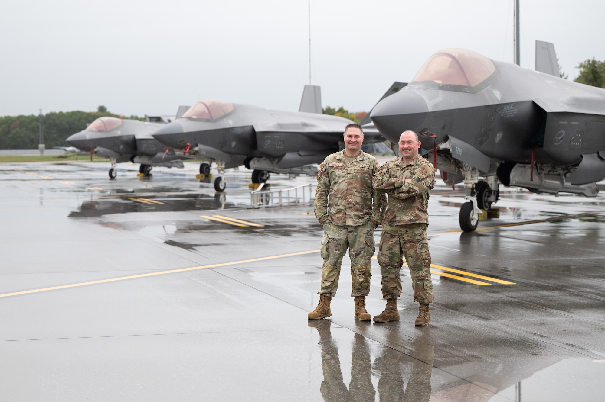 Two U.S. Air Force service members in OCP uniforms stand confidently with their arms crossed on a wet airfield, positioned in front of the sleek, grey silhouettes of two F-35 Lightning II fighter jets.
