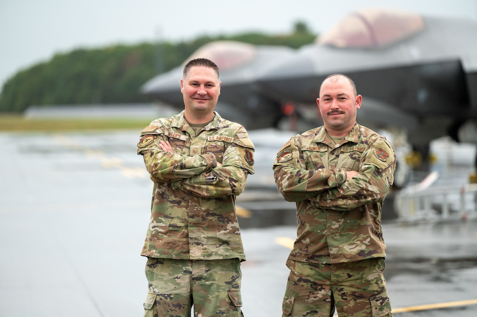 Two U.S. Air Force service members in OCP uniforms stand confidently with their arms crossed on a wet airfield, positioned in front of the sleek, grey silhouettes of two F-35 Lightning II fighter jets.