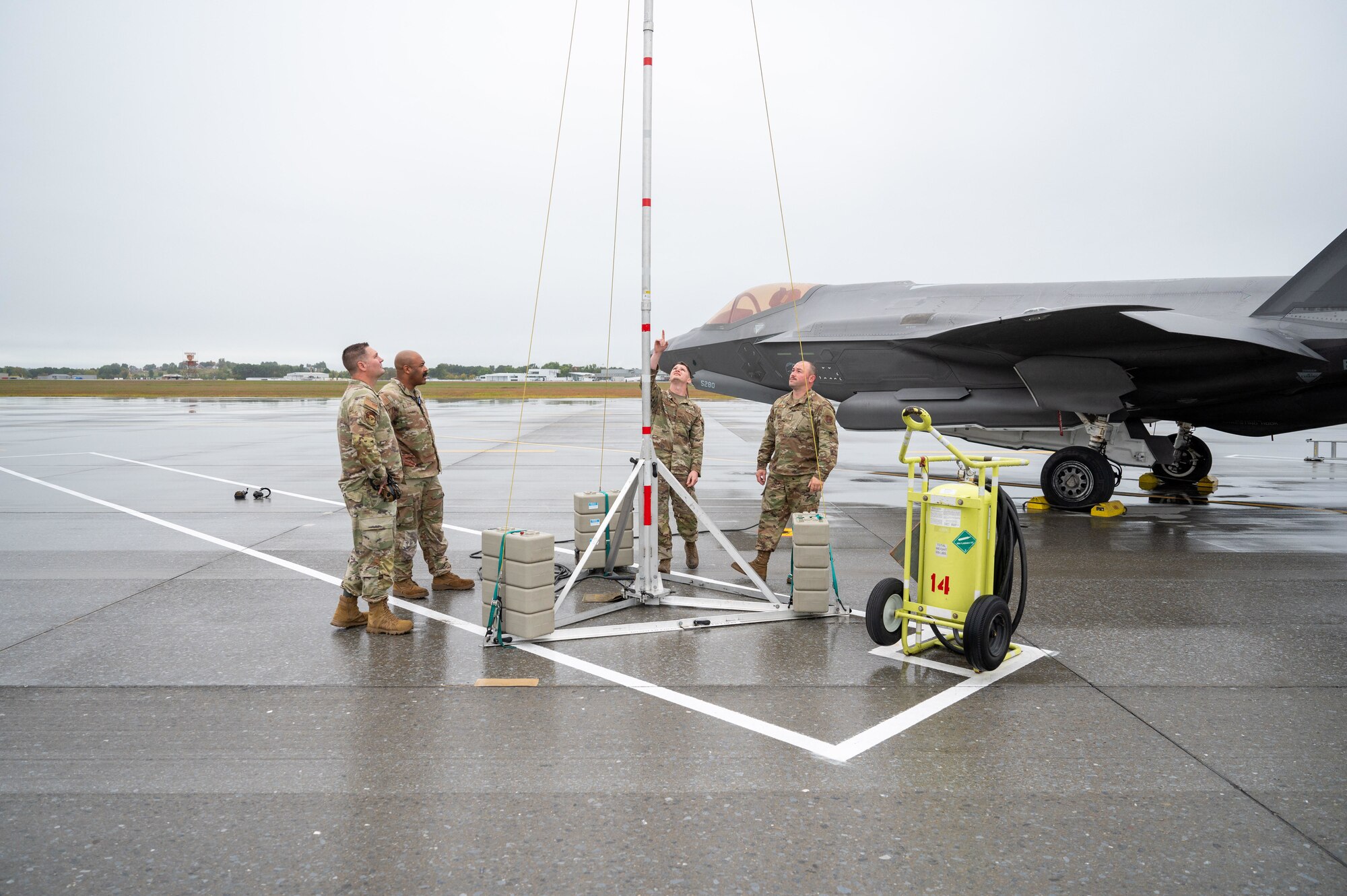 Four Air Force service members in OCP uniforms work around a lightning protection system on a wet flightline, with a stealthy F-35 Lightning II aircraft parked in the background.