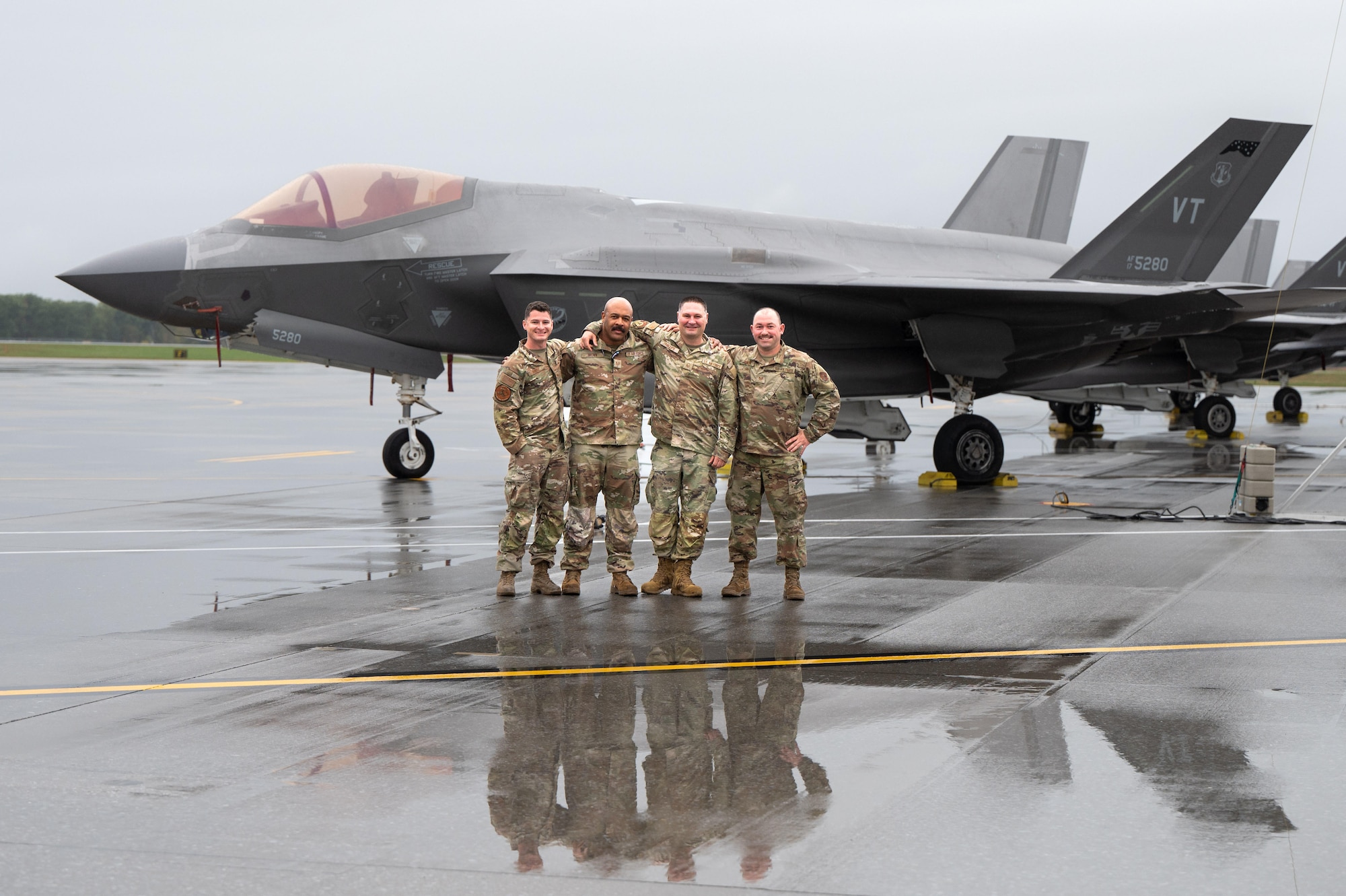 Four U.S. Air Force service members in OCP uniforms stand with their arms around each other on a wet flightline, posing in front of an F-35 Lightning II stealth fighter jet.