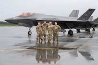 Four U.S. Air Force service members in OCP uniforms stand with their arms around each other on a wet flightline, posing in front of an F-35 Lightning II stealth fighter jet.