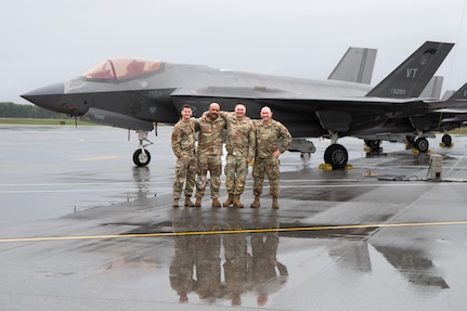 Four U.S. Air Force service members in OCP uniforms stand with their arms around each other on a wet flightline, posing in front of an F-35 Lightning II stealth fighter jet.