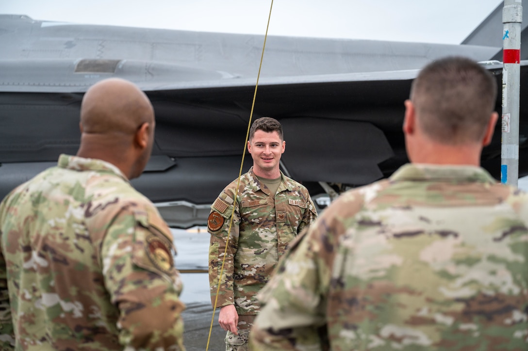 An Air Force service member in uniform smiles while speaking with two colleagues, whose backs are to the camera, in front of an F-35 Lightning II aircraft.