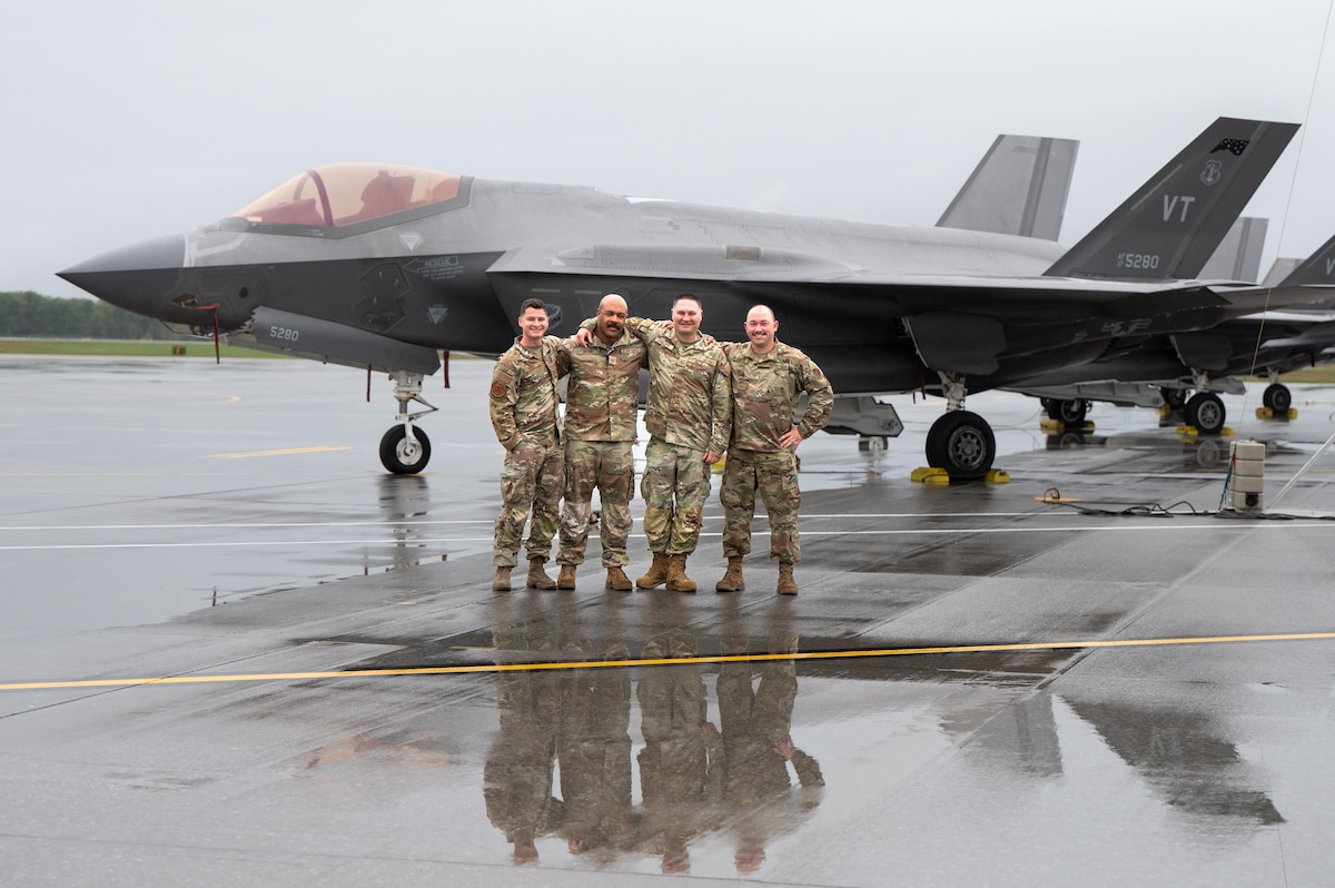 Four U.S. Air Force service members in OCP uniforms stand with their arms around each other on a wet flightline, posing in front of an F-35 Lightning II stealth fighter jet.