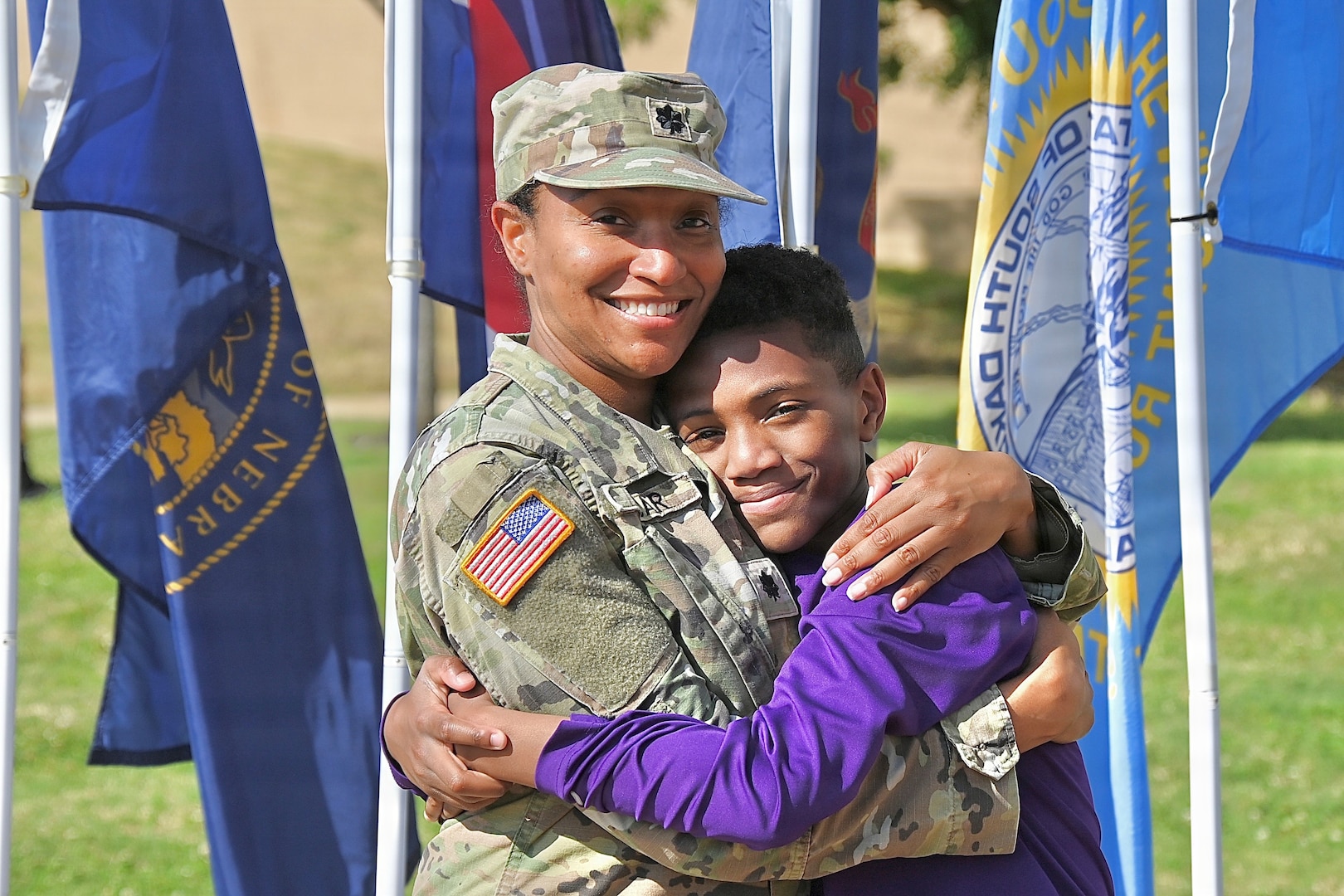 Military mom hugs her son in front of flags.