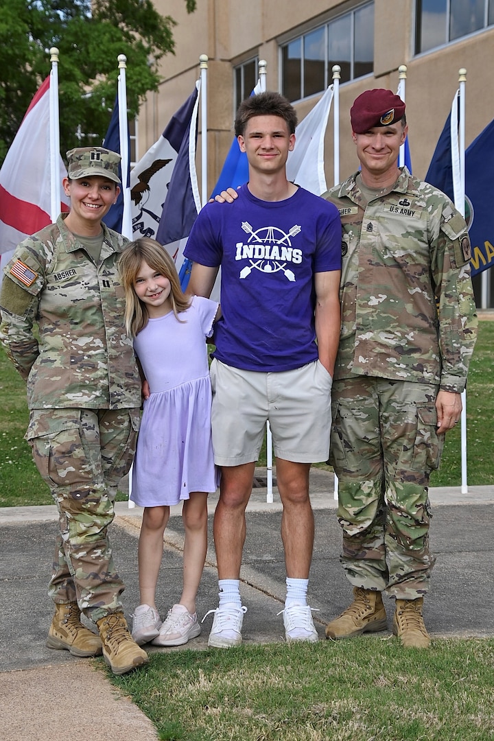 Soldier parents pose with their two children in front of a line of flags.