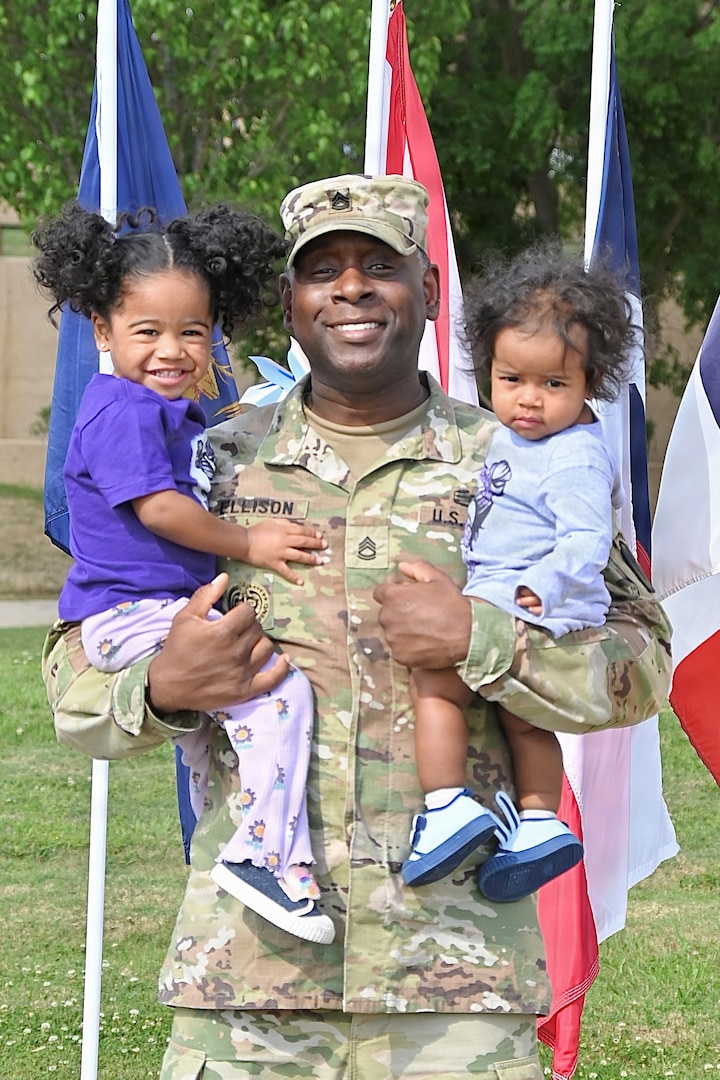 Soldier father holding a daughter in each arm standing in front of flags.