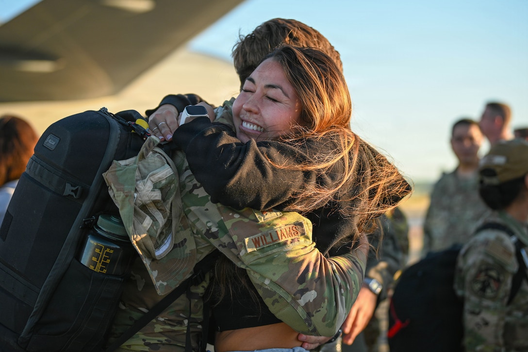 A civilian smiles while hugging an airman wearing a backpack on a flight line.