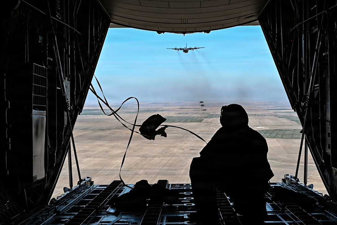 An airman, shown from behind, crouches at the back of an open, in-flight aircraft facing packages descending near another plane.