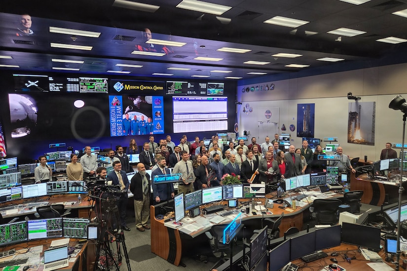 Dozens of people in business attire pose for a photo inside a workstation with multiple computers on surrounding desks and large screens in the background. There are photos on the walls of spacecraft launching into space.
