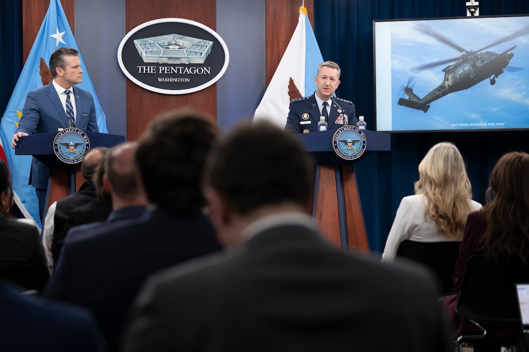 The Joint Chiefs chairman speaks at a lectern by a screen displaying a helicopter image; The secretary of war stands at another lectern.