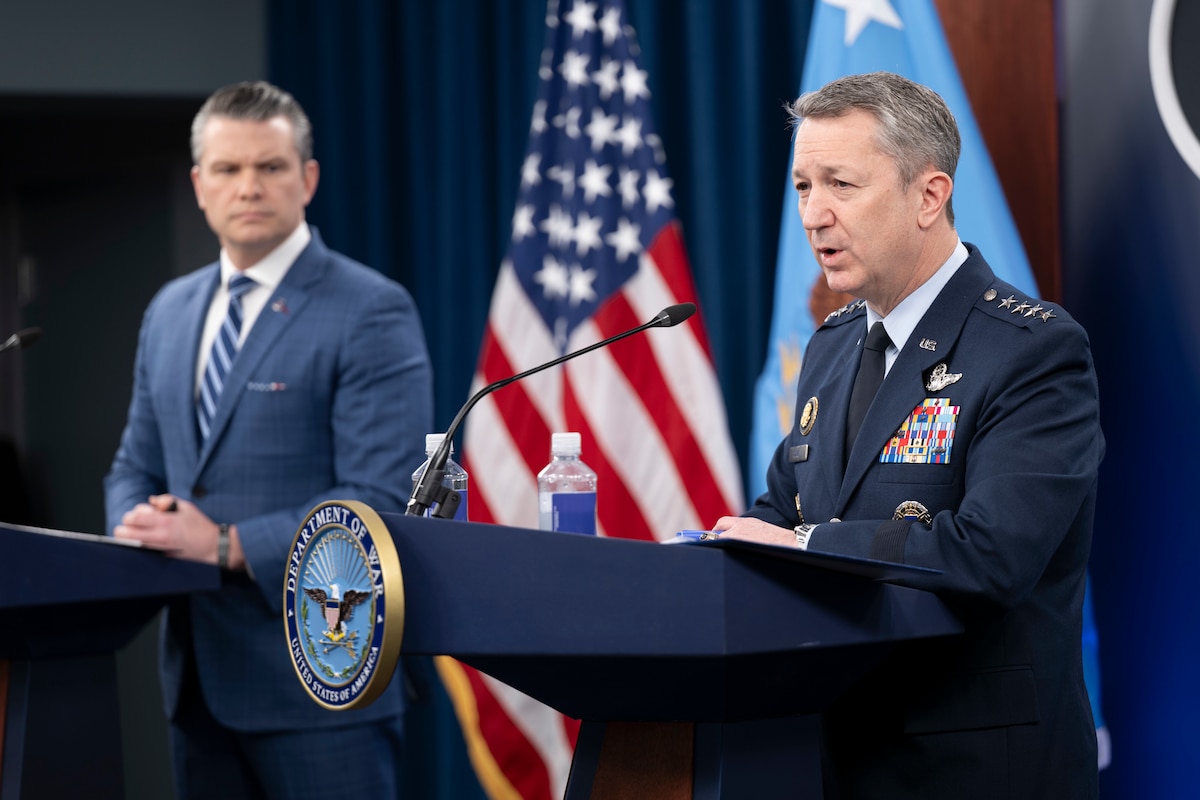A man in business attire and another in a formal military uniform stand behind lecterns and speak to an audience. Behind them is an American flag.