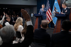 A man in business attire and another in a formal military uniform stand behind lecterns and answer questions from an audience. Behind them is an American flag.