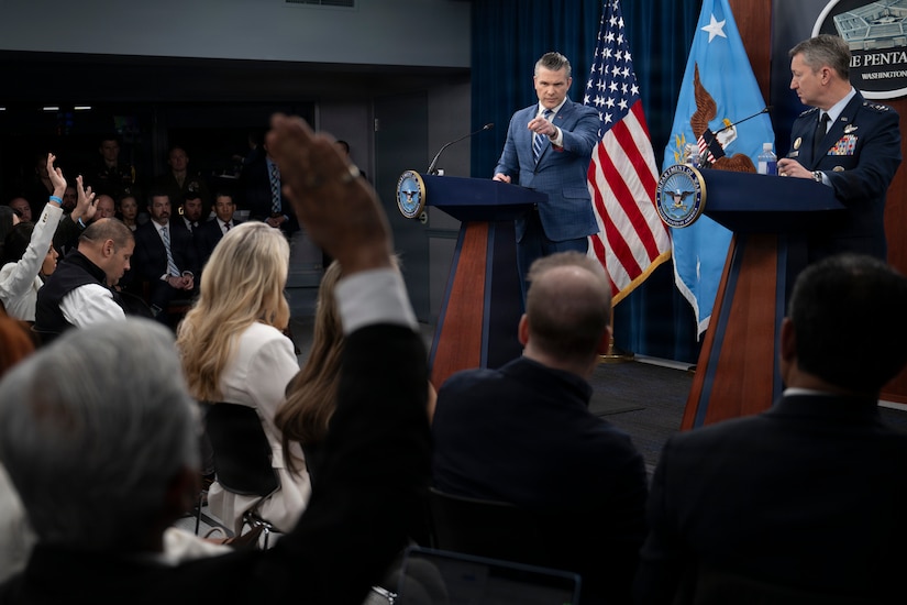 A man in business attire and another in a formal military uniform stand behind lecterns and answer questions from an audience. Behind them is an American flag.