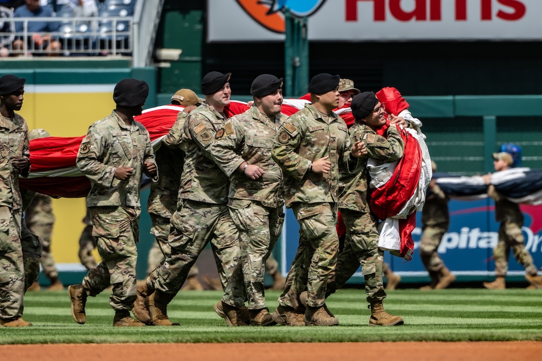 Service members hold U.S. flag on baseball field.