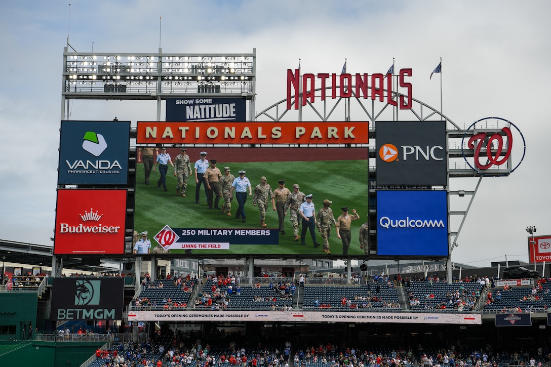 Service members hold U.S. flag on baseball field.