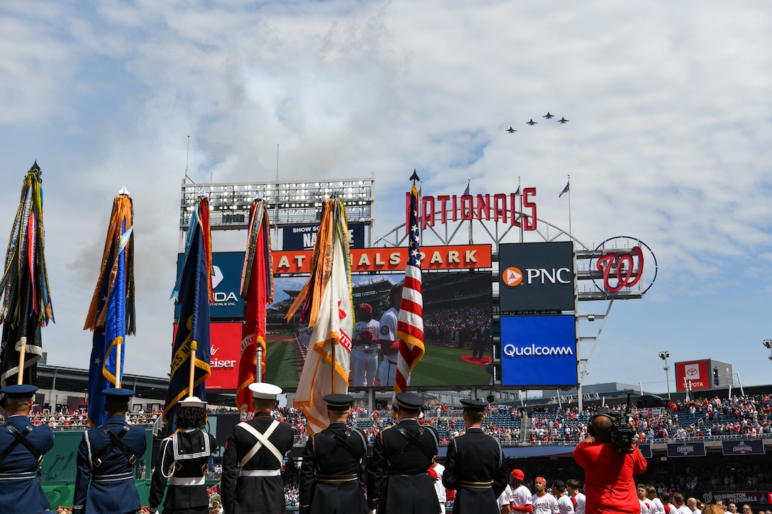 Service members hold U.S. flag on baseball field.
