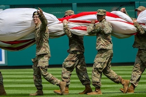 Service members hold U.S. flag on baseball field.