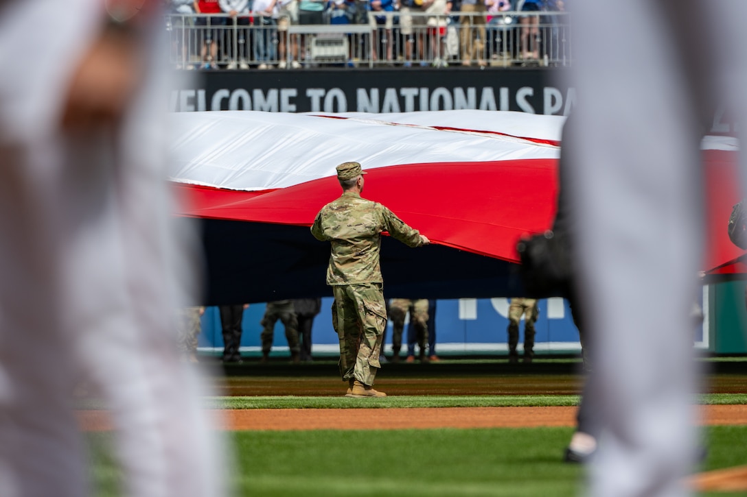 Service members hold U.S. flag on baseball field.
