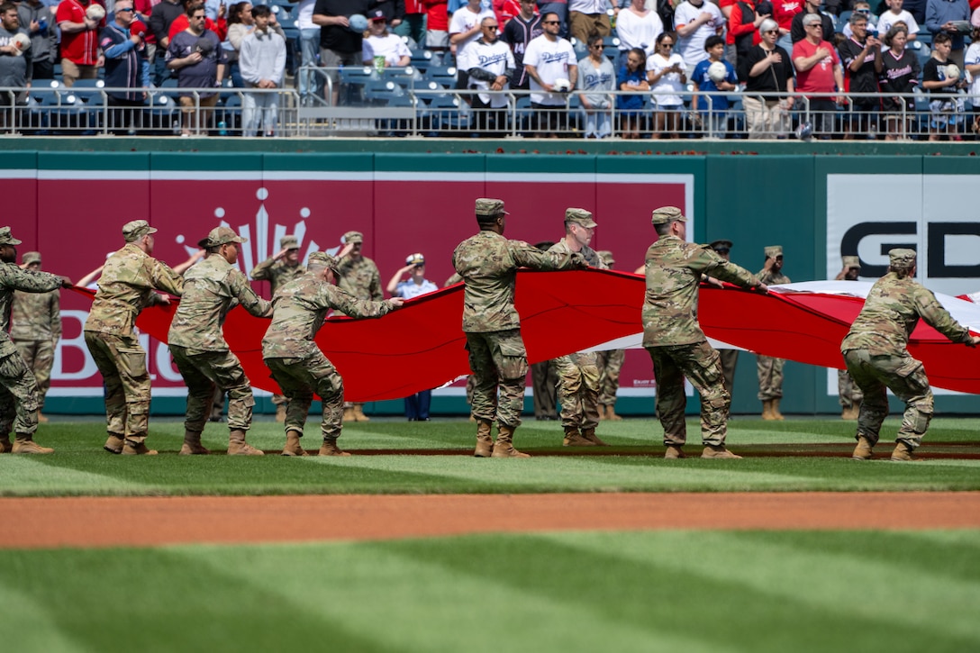 Service members hold U.S. flag on baseball field.