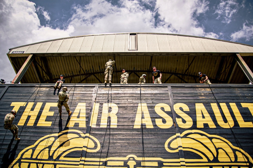Two soldiers hold on to ropes and descend a wall with the words “The Air Assault” painted on it in golden yellow, as others watch from the top.