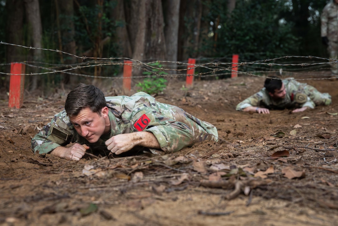 Two soldiers in camouflage uniforms lay on dirt under barbed wire with their heads slightly raised.
