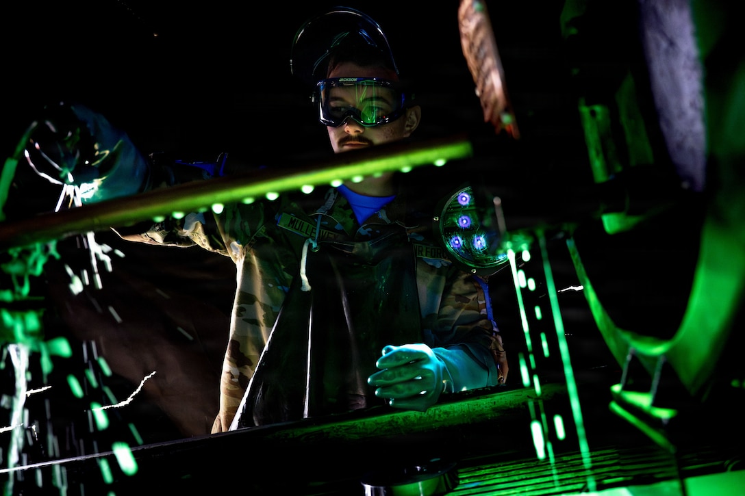 A service member wearing goggles and rubber gloves works on equipment in a dark space illuminated by green light.