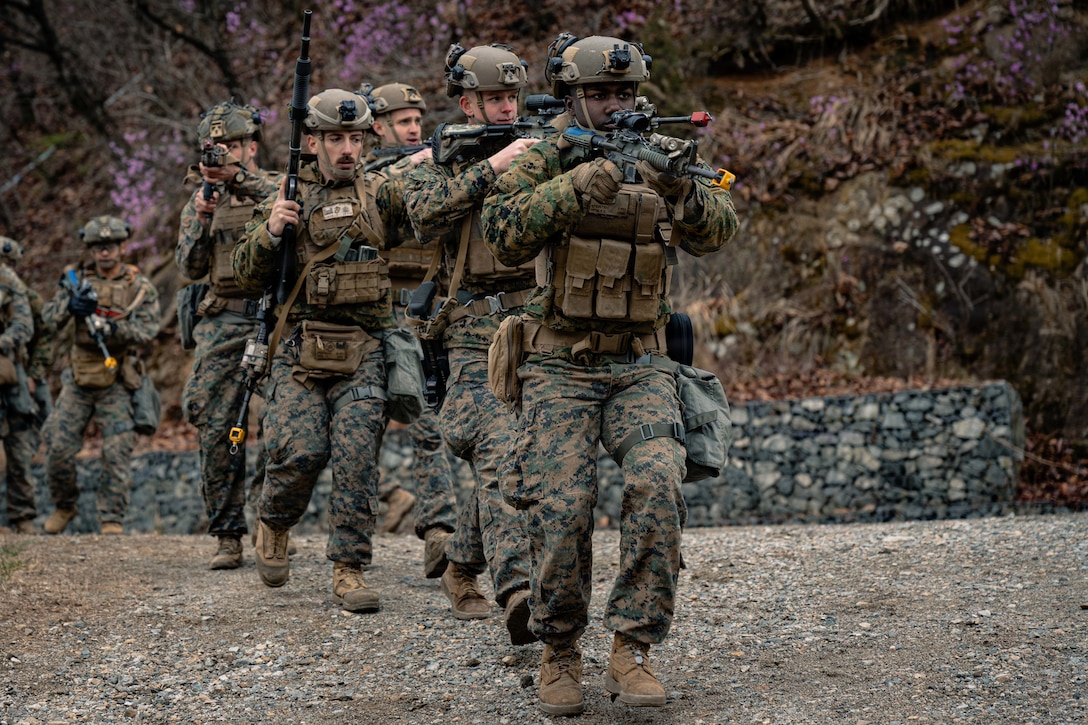 Marines in camouflage combat gear and carrying rifles advance in a single-file row in a rocky, hilly area during daytime, as other Marines watch in the background.