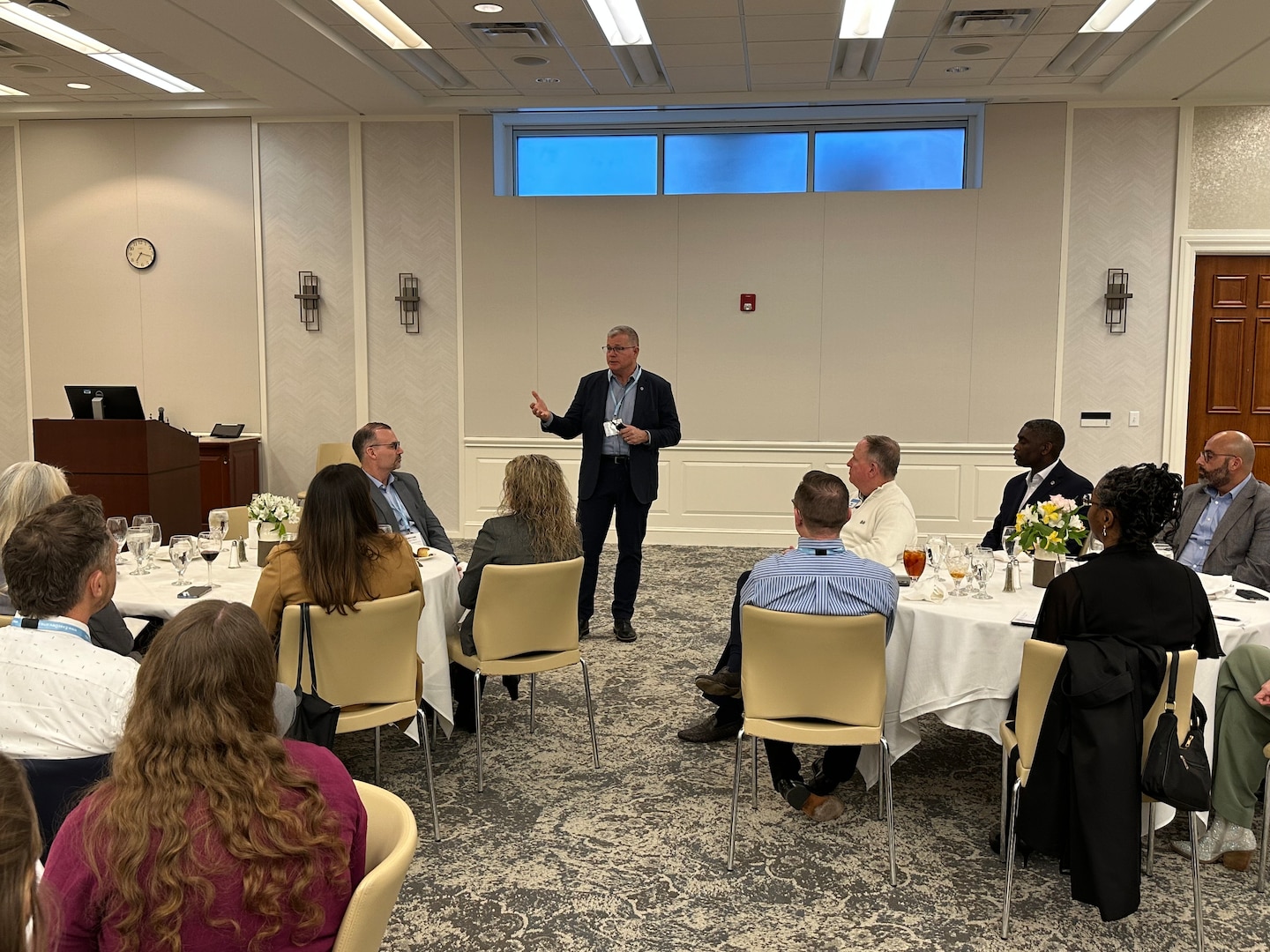 A man stands at the front of a banquet room, speaking to tables of people sitting in front of him